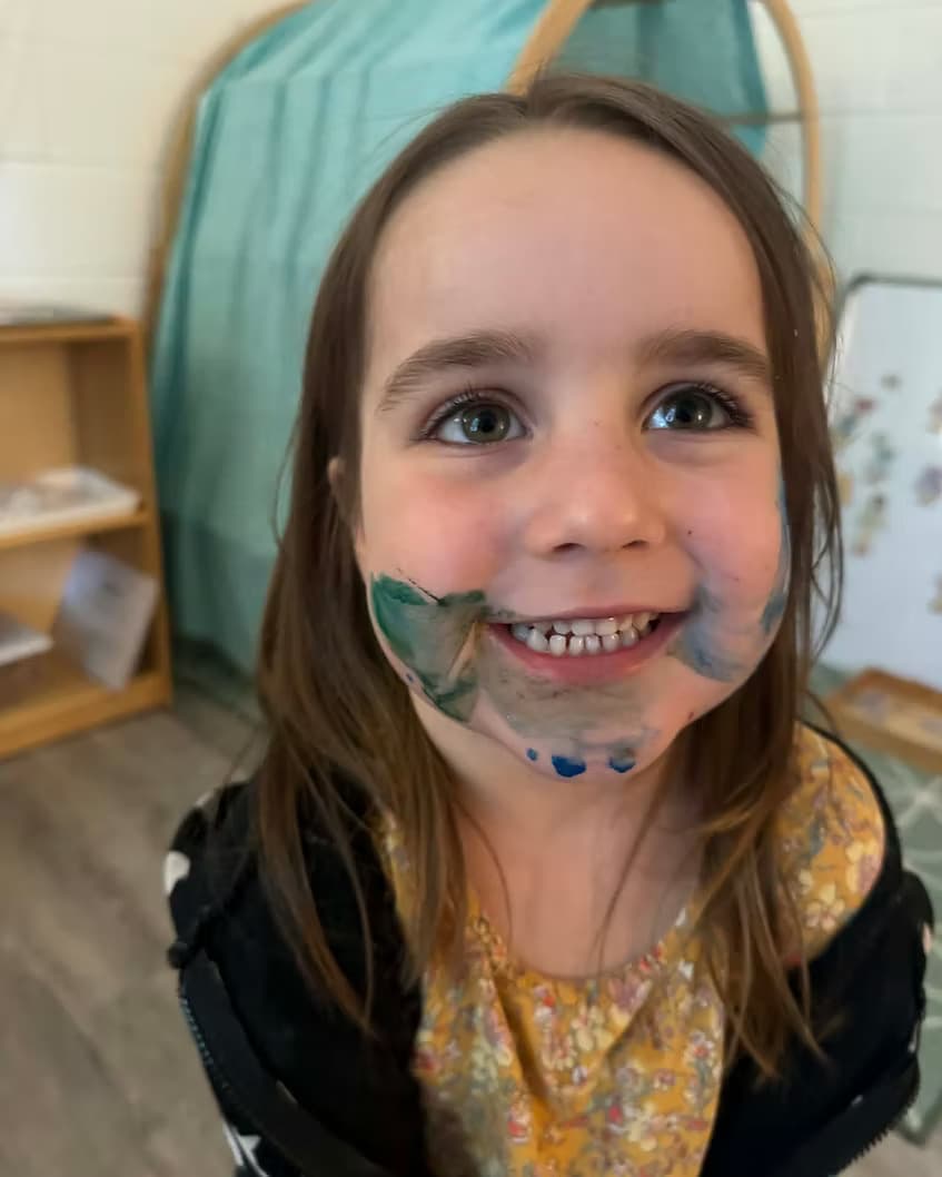 A preschooler with paint on their face, smiling during an art activity